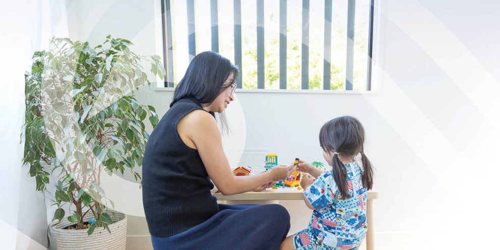 Image of Ying sitting at a small table with a young one playing with toys.